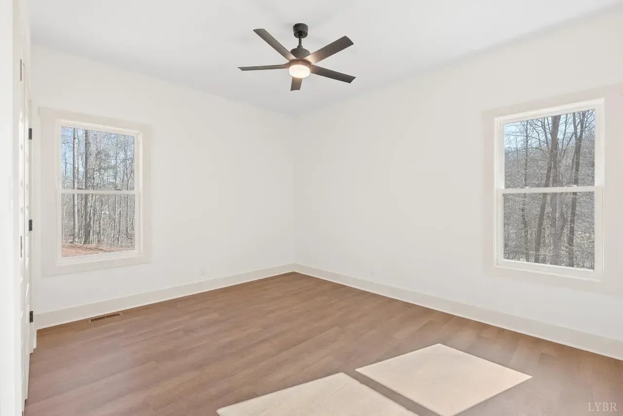 An empty bedroom with light wood flooring, white walls, two windows overlooking trees, and a ceiling fan.