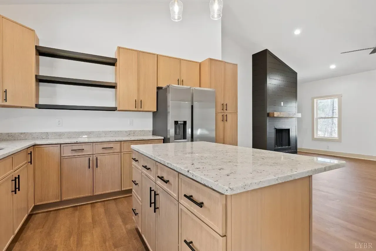 A modern kitchen with light wood cabinets, granite countertops, a central island, and an adjacent fireplace feature wall.