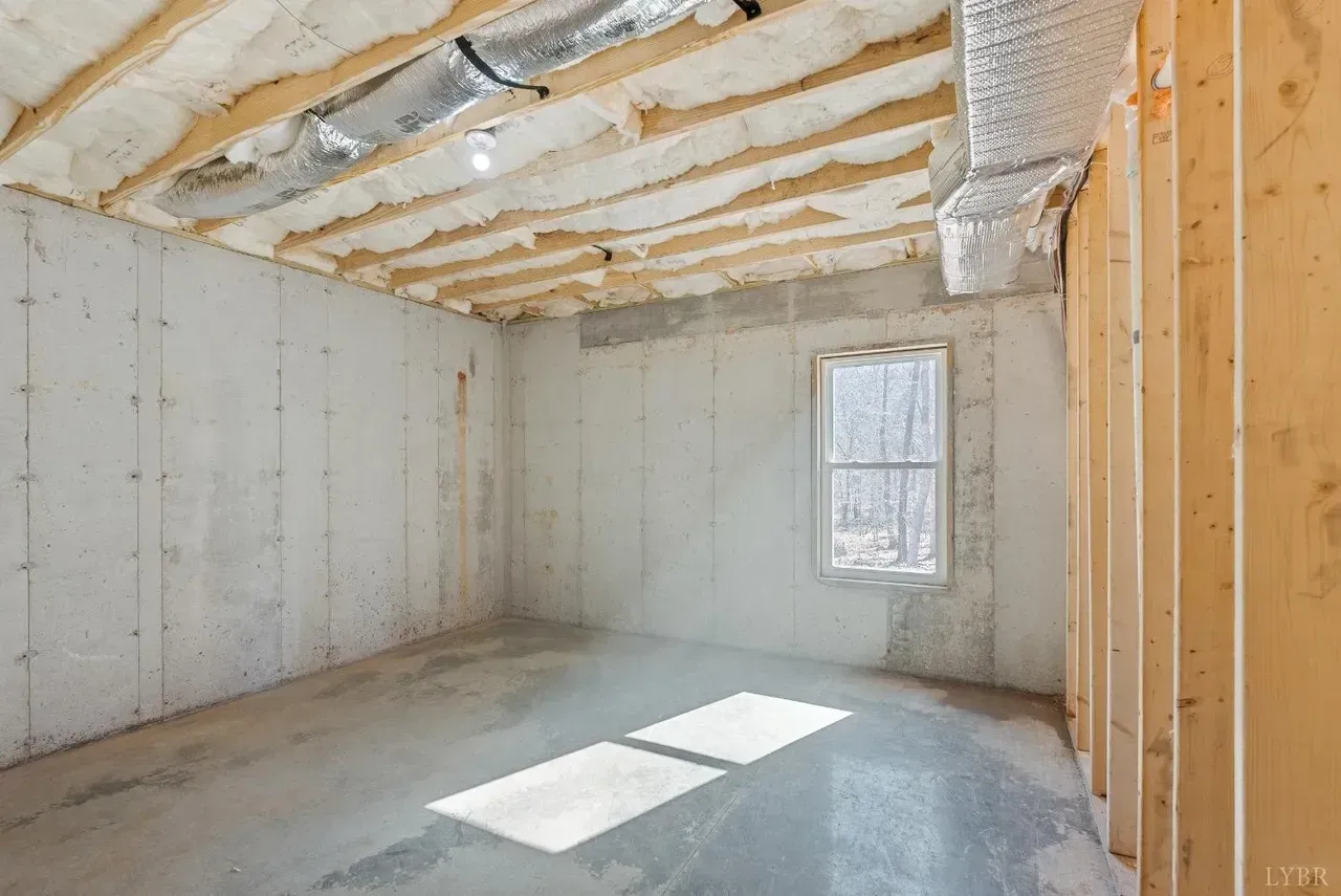 An unfinished basement with concrete walls, exposed wooden ceiling joists with insulation, and a single window.