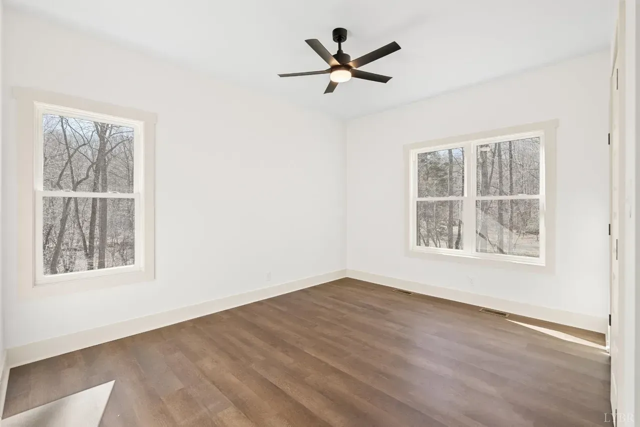 An empty room with white walls, wood-look flooring, two windows looking onto trees, and a ceiling fan.