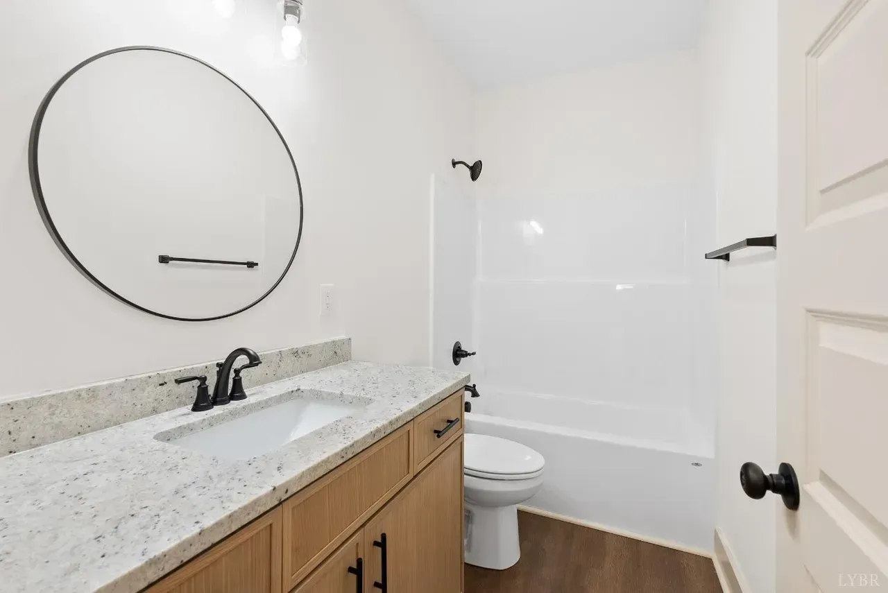 A bathroom with a light wood vanity, speckled granite countertop, round mirror, white bathtub, and toilet.