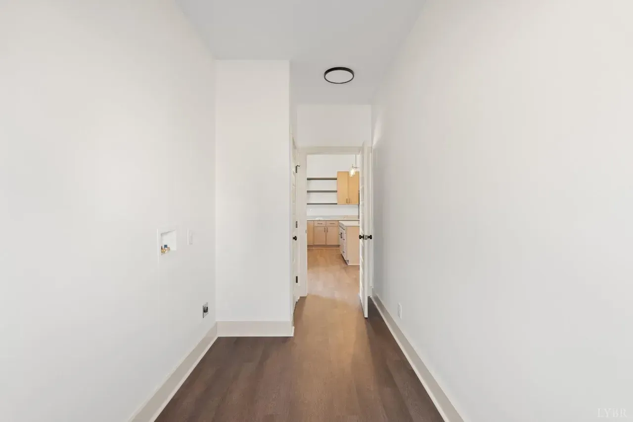 A narrow, white hallway with wood-tone flooring leading to an open doorway that shows a brightly lit kitchen.