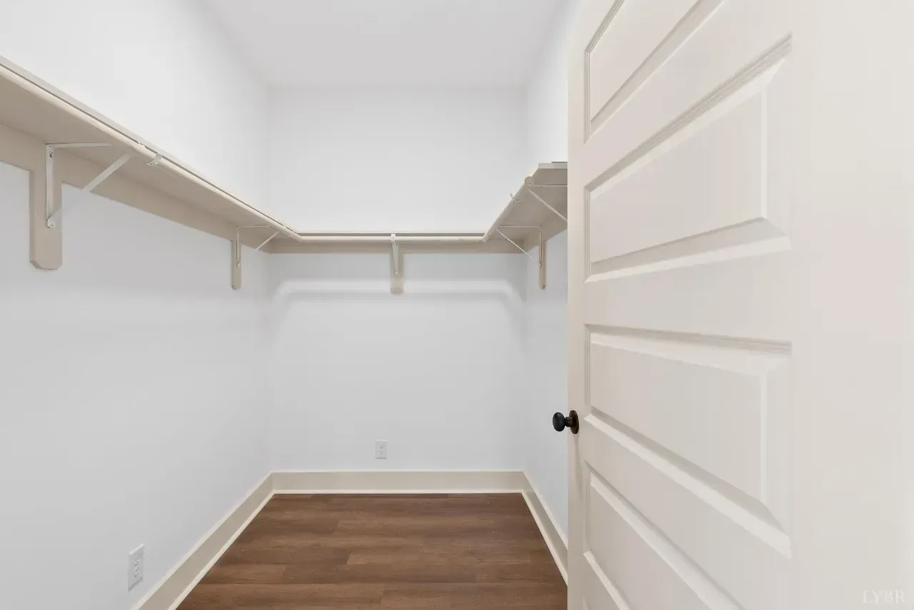An empty walk-in closet with white walls, wood-look flooring, and a wrap-around shelving unit, viewed through a doorway.