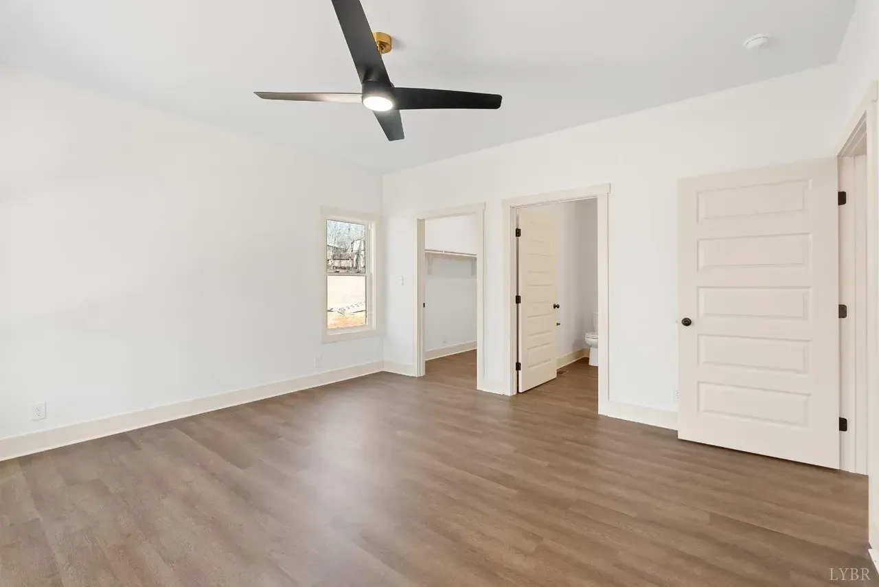 An empty bedroom with white walls, wood-look flooring, a ceiling fan, a window, and two open doorways.