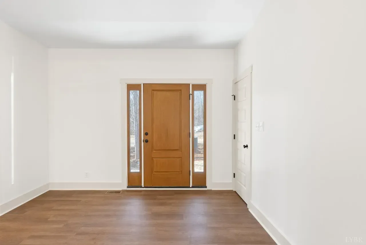 An interior view of a room with light wood floors, white walls, a wooden front door with side windows, and a white door.
