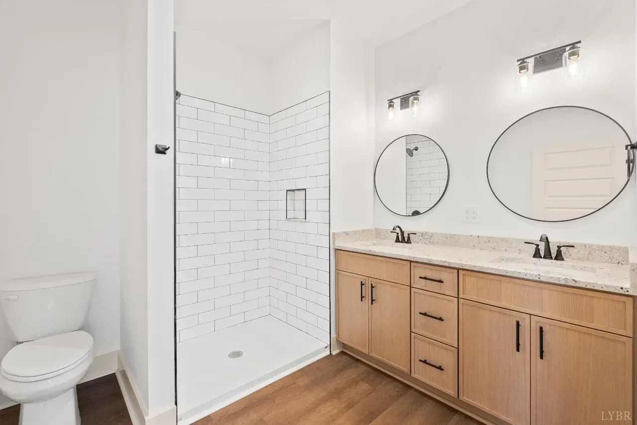 Modern bathroom with light wood vanity, dual sinks, oval mirrors, white subway tile shower, and white toilet.