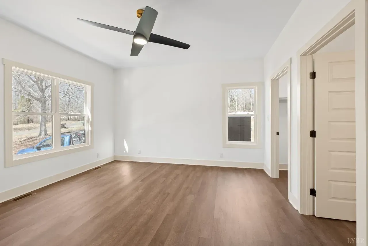 An empty bedroom with white walls, wood-look flooring, a ceiling fan, two windows, and an open doorway to a closet.