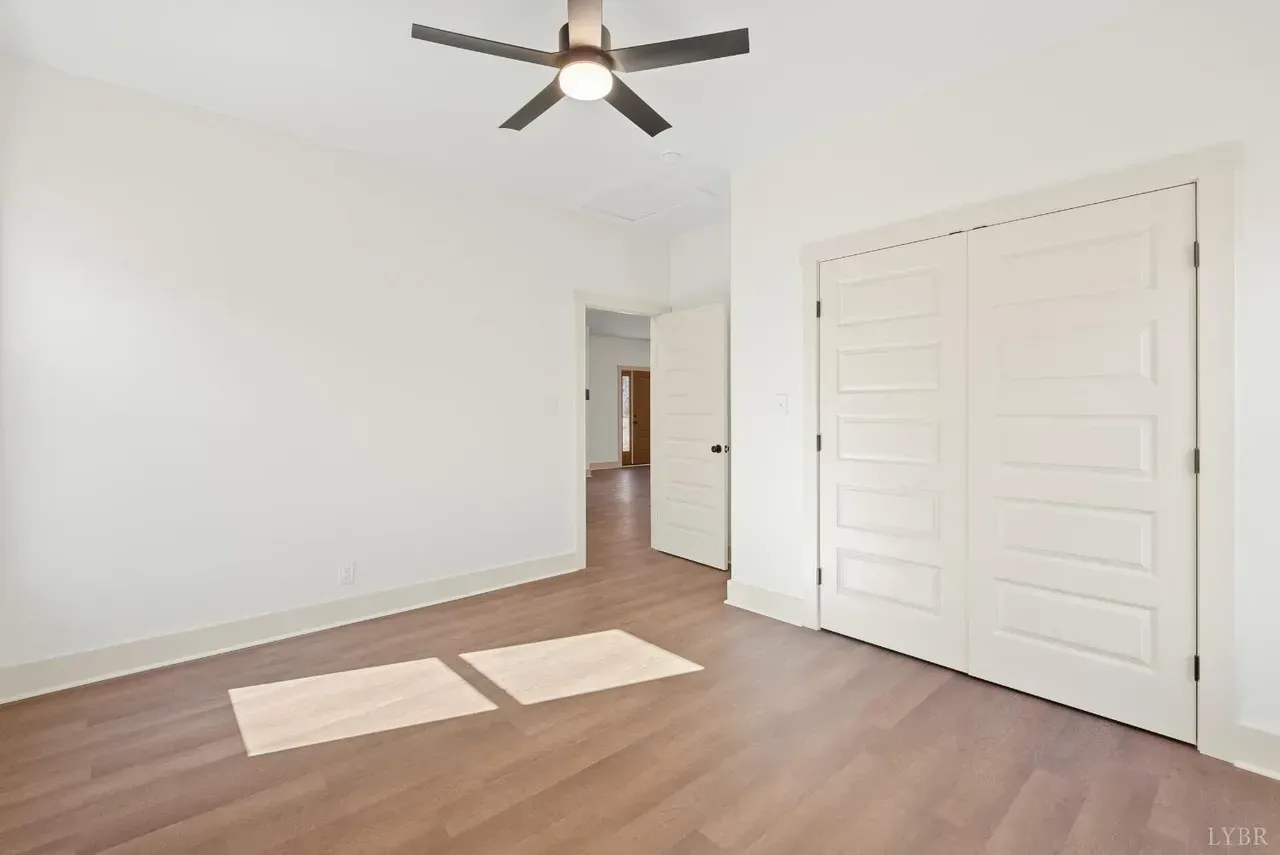A white-walled room with wood-look flooring, a ceiling fan, a large closet with paneled doors, and an open doorway.