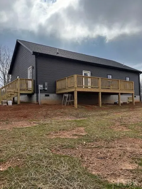 A dark gray house with a wooden deck and staircase, set on a rural lot with patches of grass and bare red soil.
