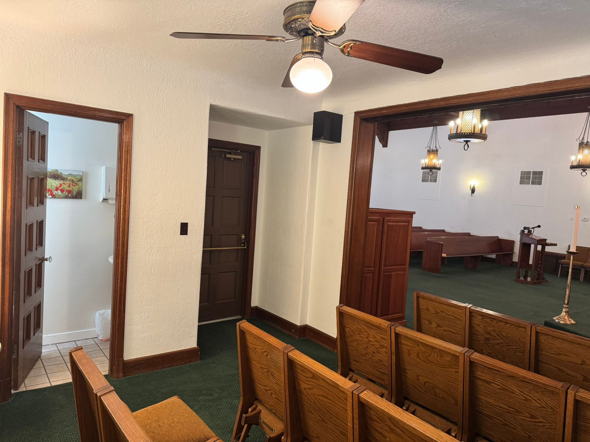 Interior of a chapel with pews, doorway to a restroom, and a doorway to a larger space with more seating.
