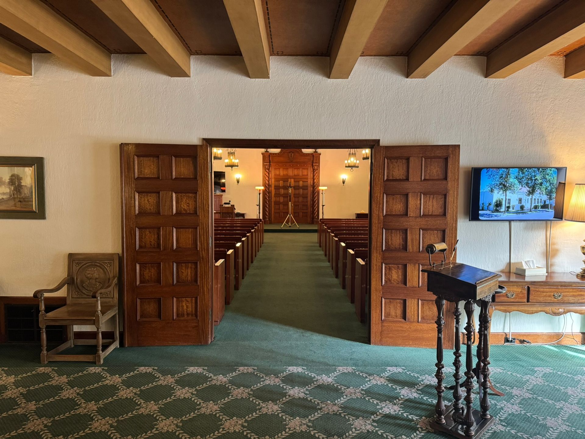 Interior of a chapel with open wooden doors, rows of pews, and a stage. Green carpet, wood beams.