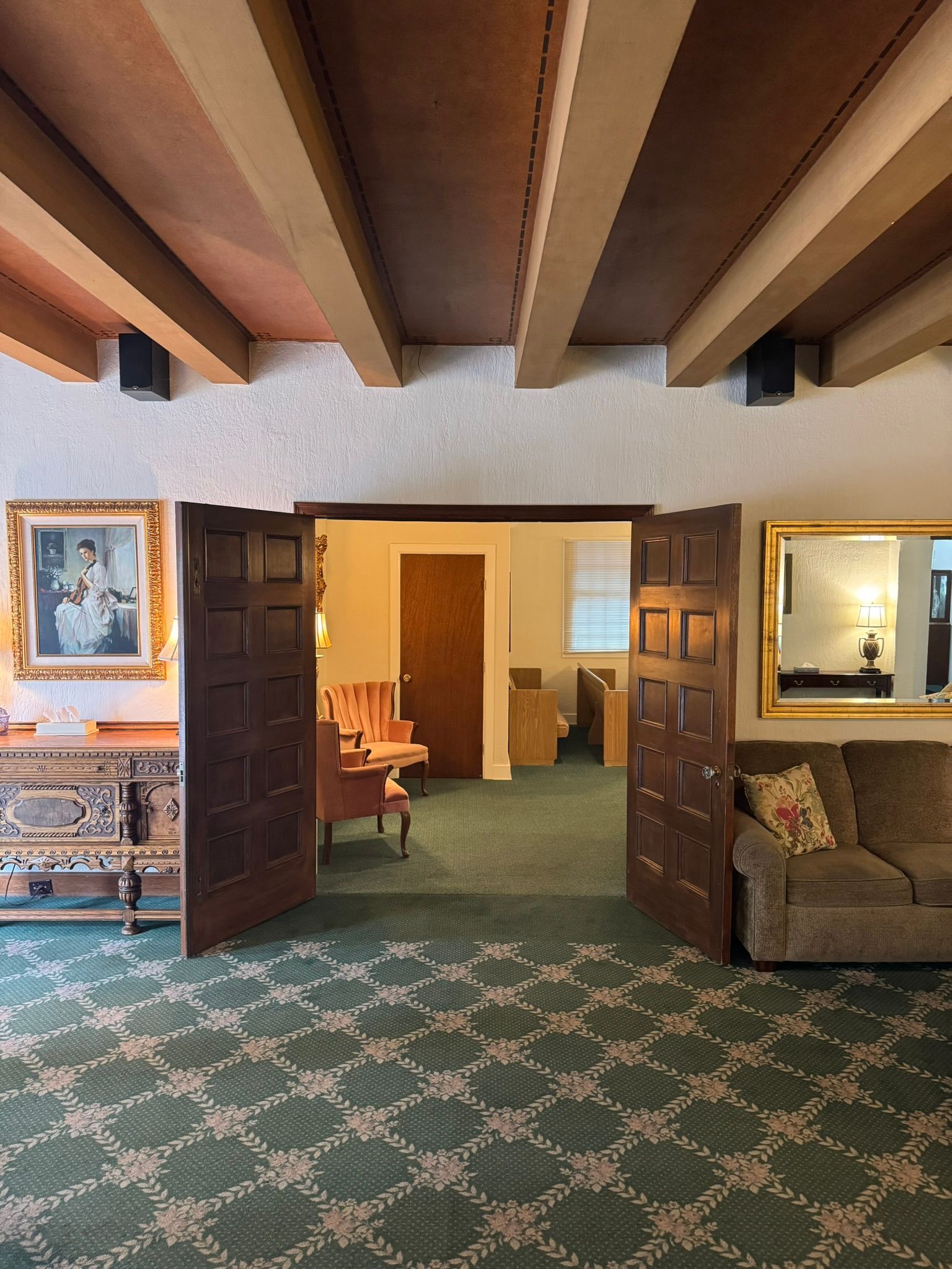 Wooden double doors open into a room with green carpet and furniture. A framed photo and mirror are visible.