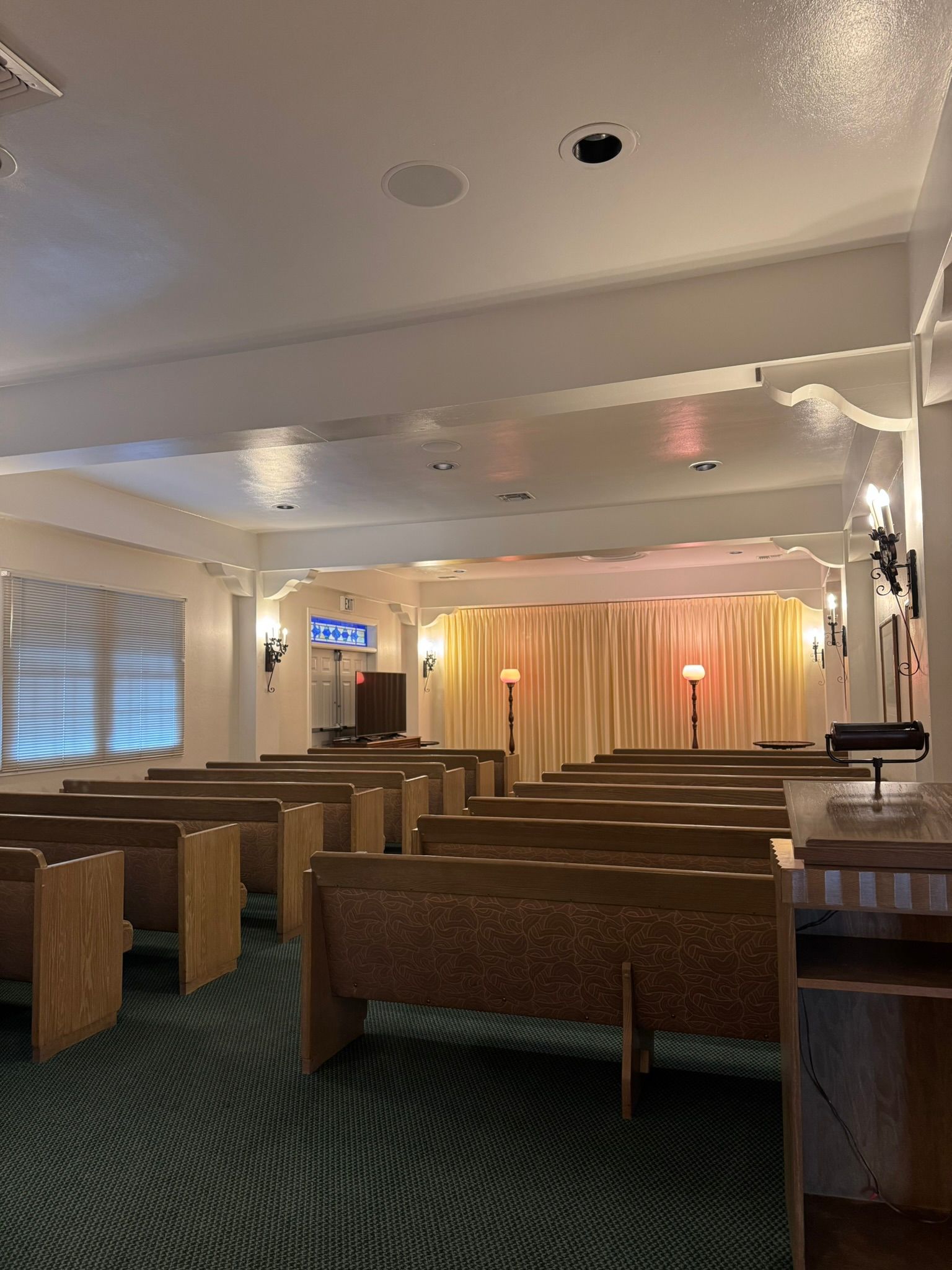 Interior of a funeral home chapel with rows of wooden pews facing a stage with curtains.