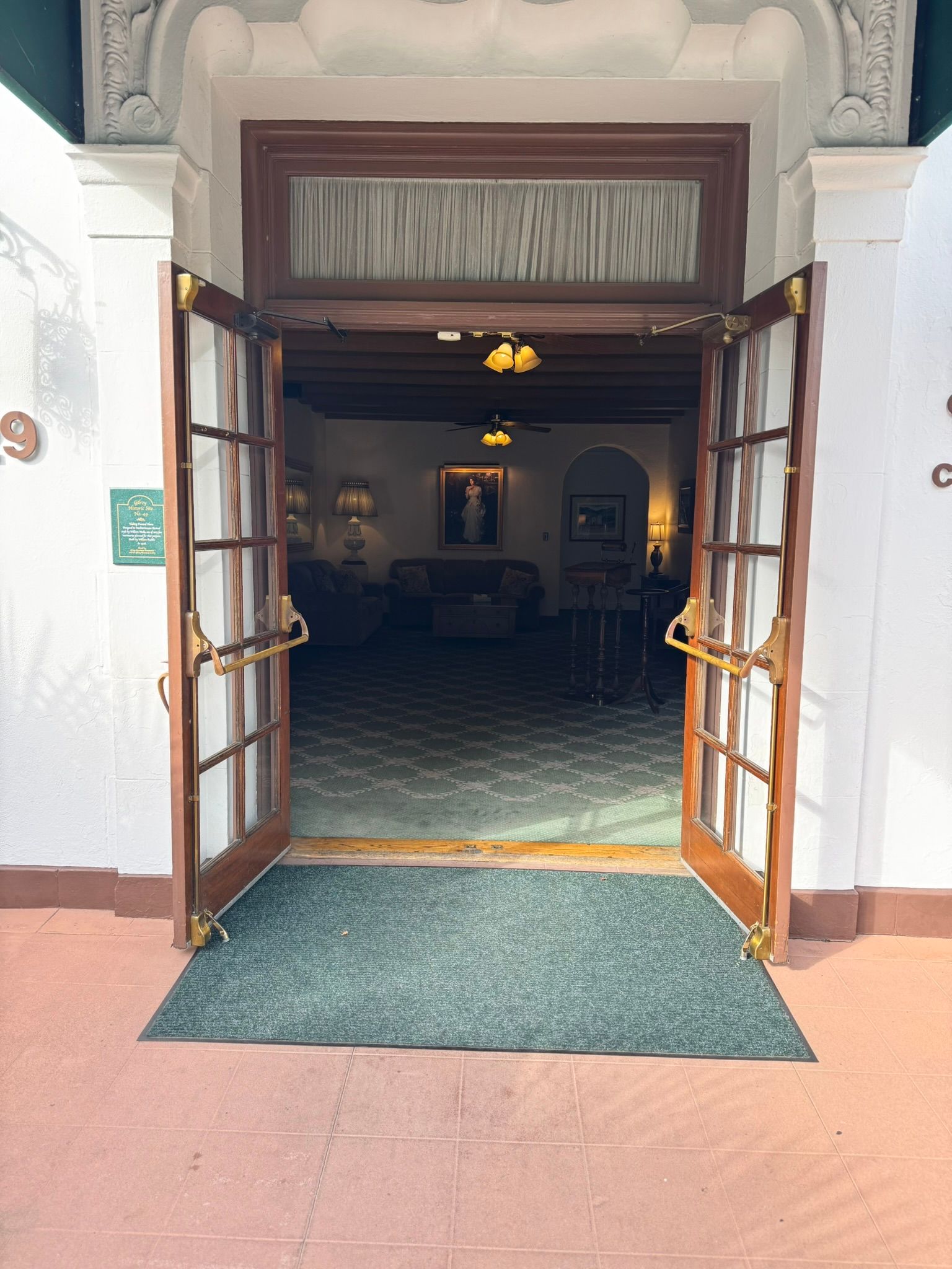 Double wooden doors open into a building lobby, a green rug on the entrance.