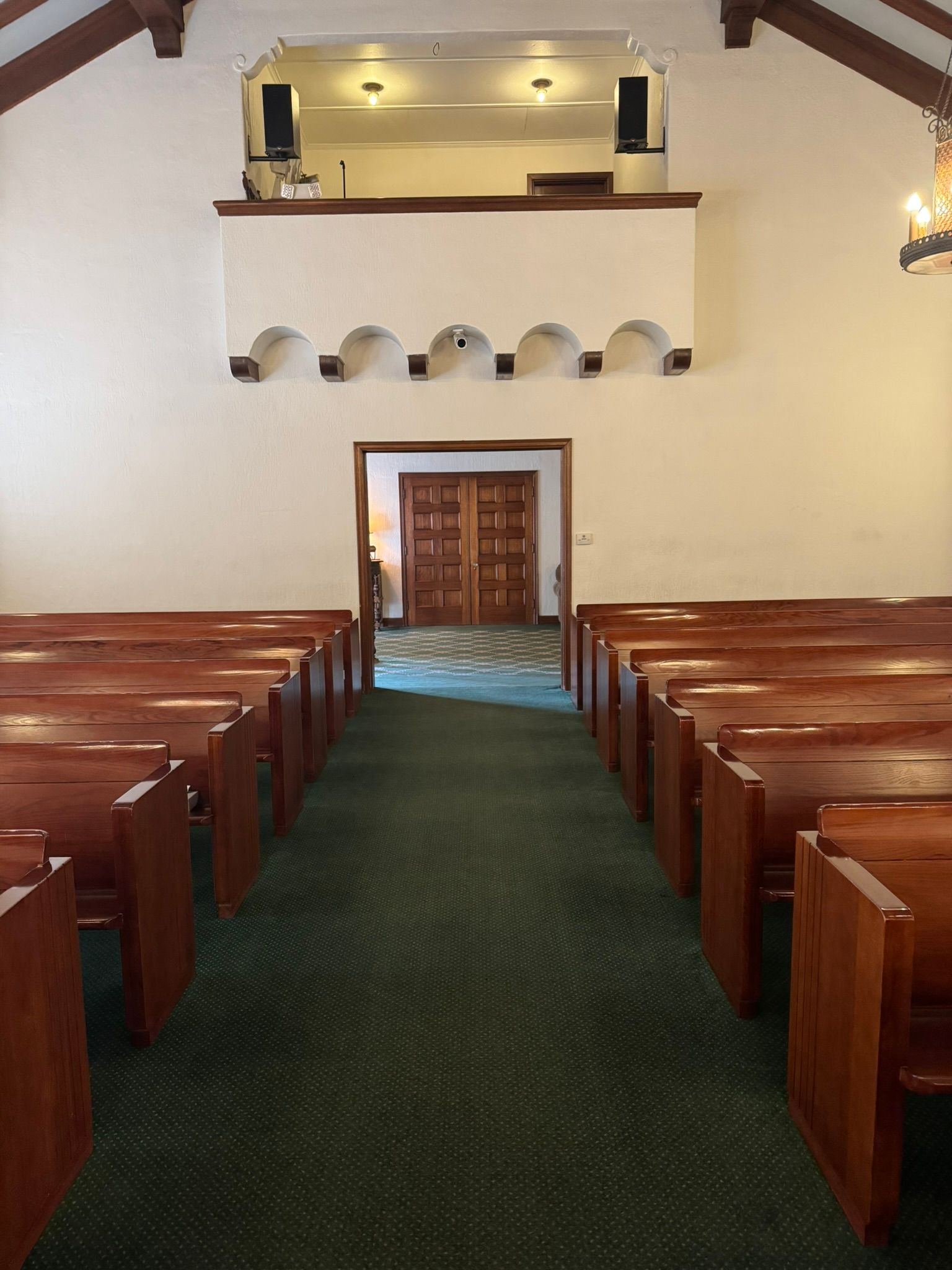 Interior of a chapel with rows of wooden pews, green carpeted aisle, and a balcony above the entrance.