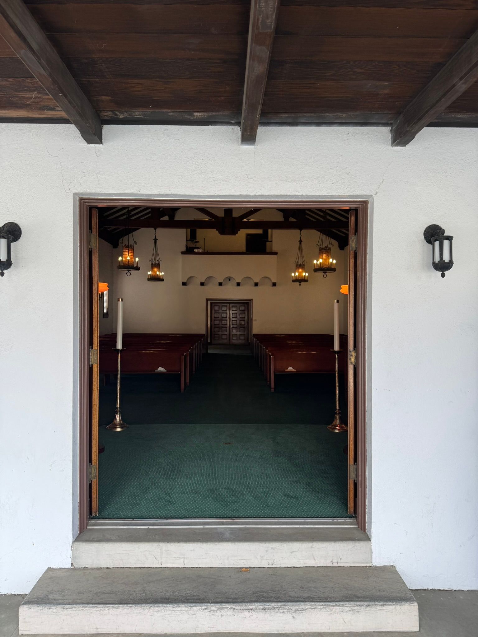 Open doorway into a chapel with green carpet, pews, and hanging lanterns. White walls and stone steps.