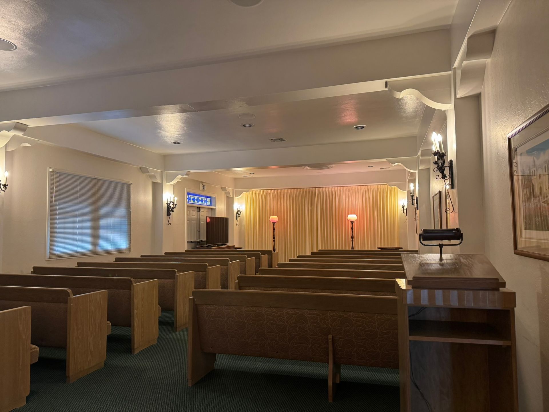 Interior of a chapel with rows of wooden pews facing a curtained stage, soft lighting.