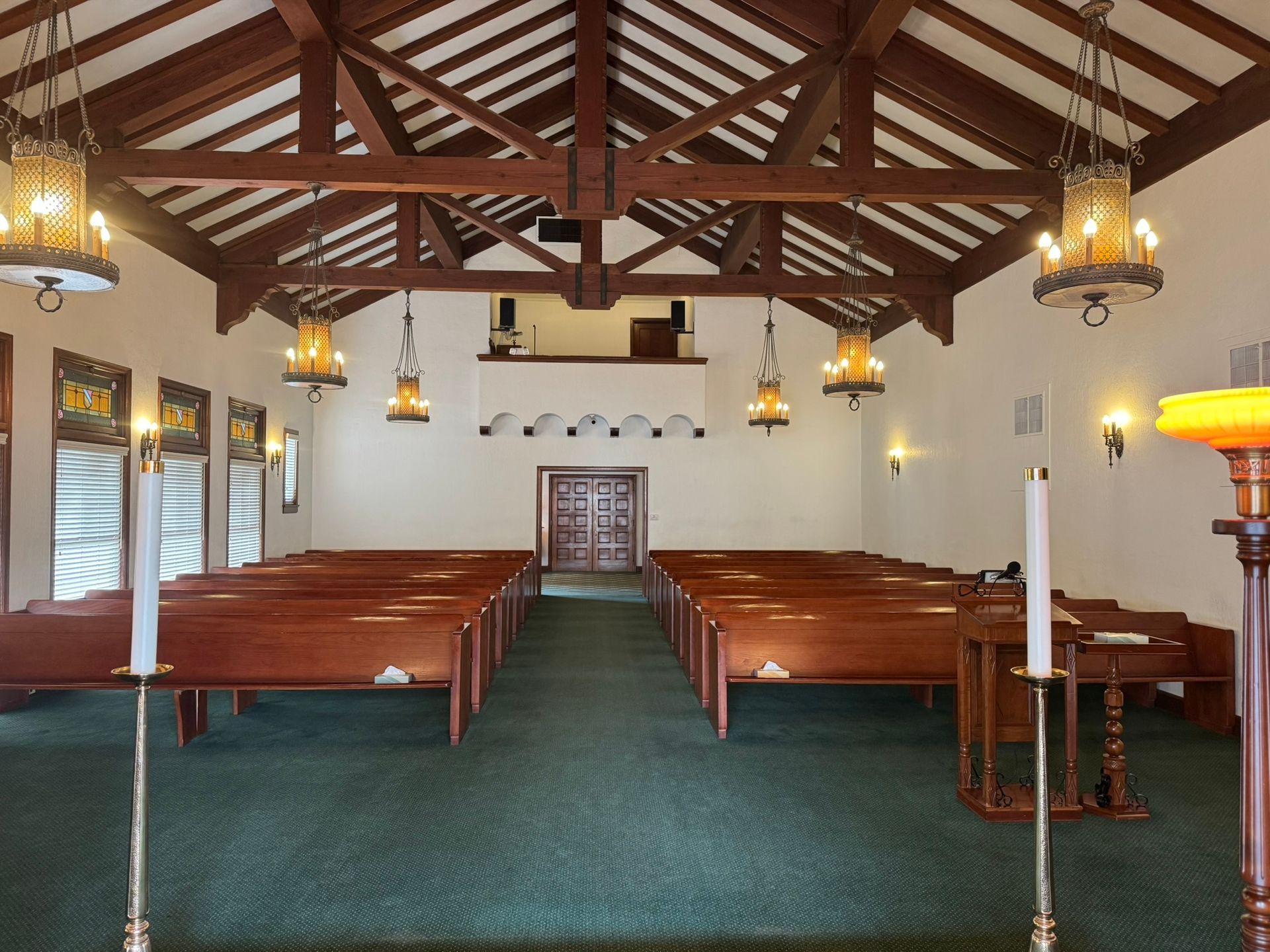Interior of a chapel with wooden pews, high ceiling, chandeliers, and a central doorway. Green carpet.