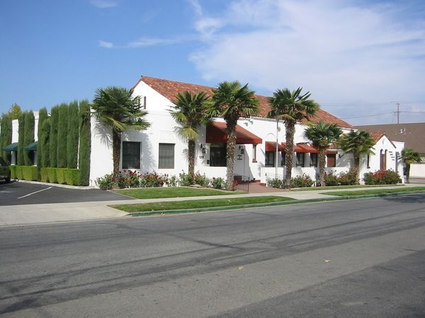 White building with red-tiled roof, palm trees, and green hedges along the sidewalk; blue sky.