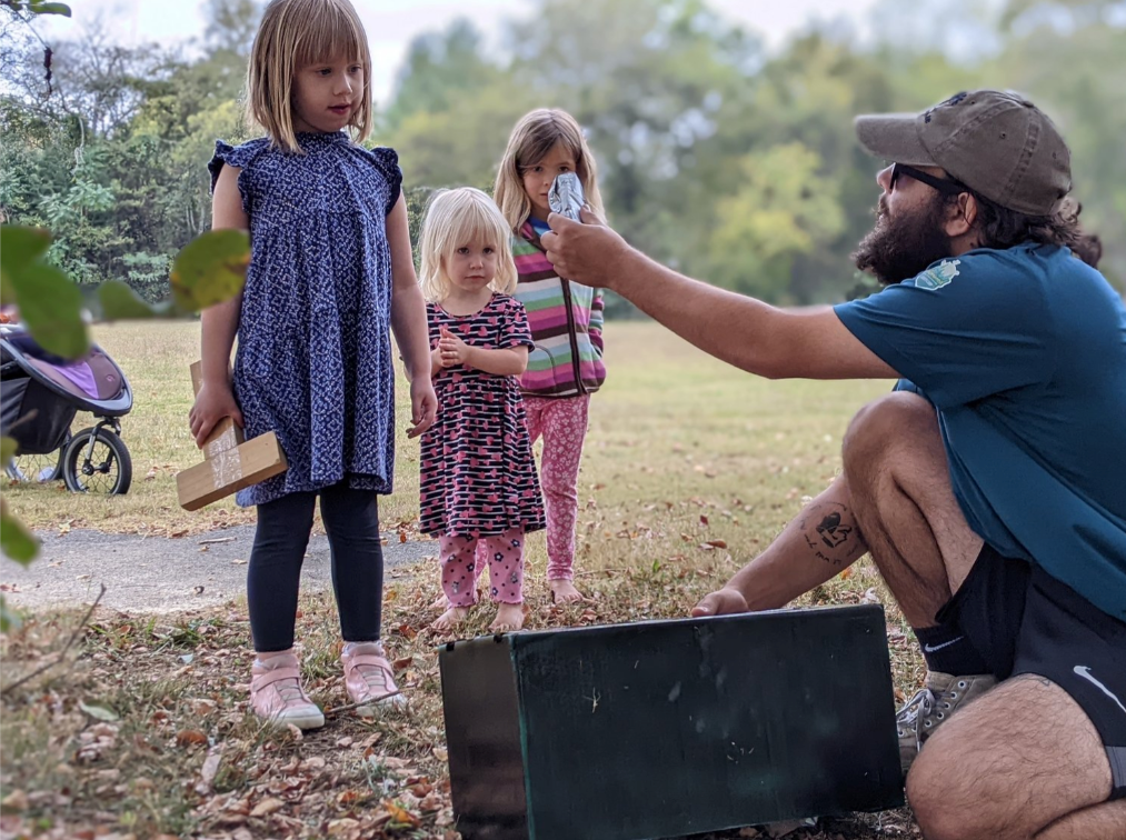 A man is kneeling down in front of two little girls and holding a light bulb.
