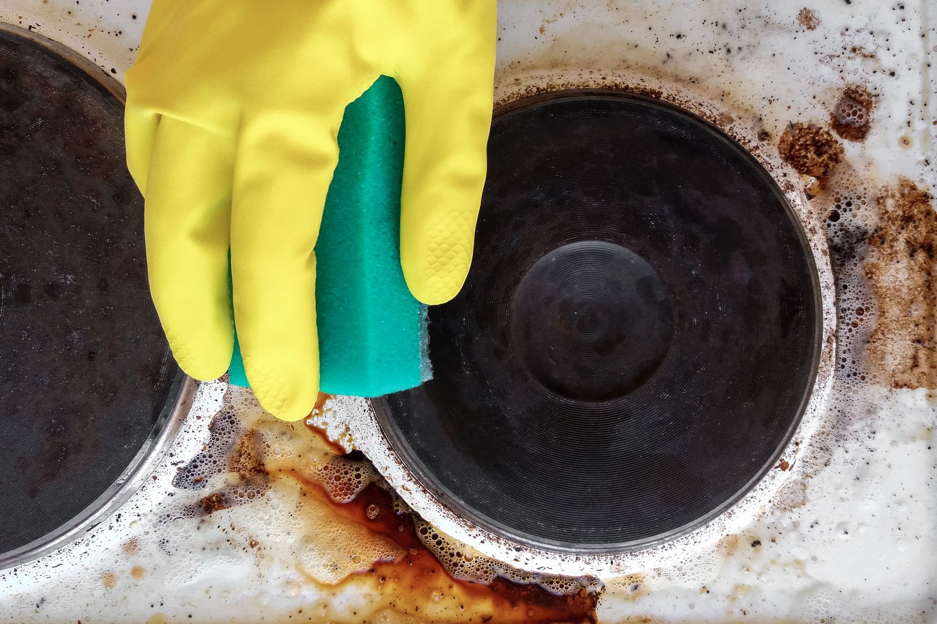 A person wearing yellow gloves is cleaning a stove top with a sponge.