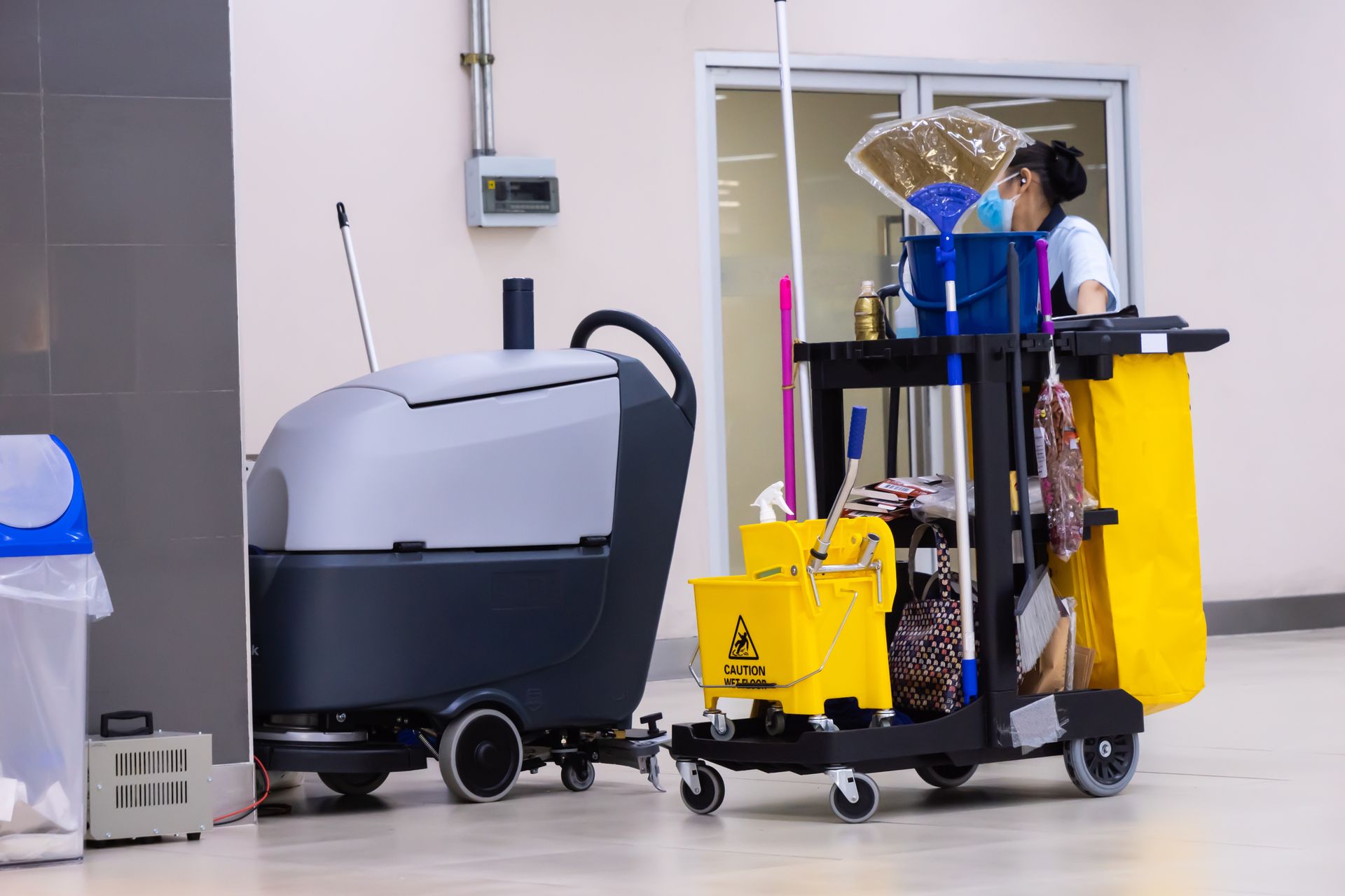 A woman is standing next to a cleaning cart in a hallway.