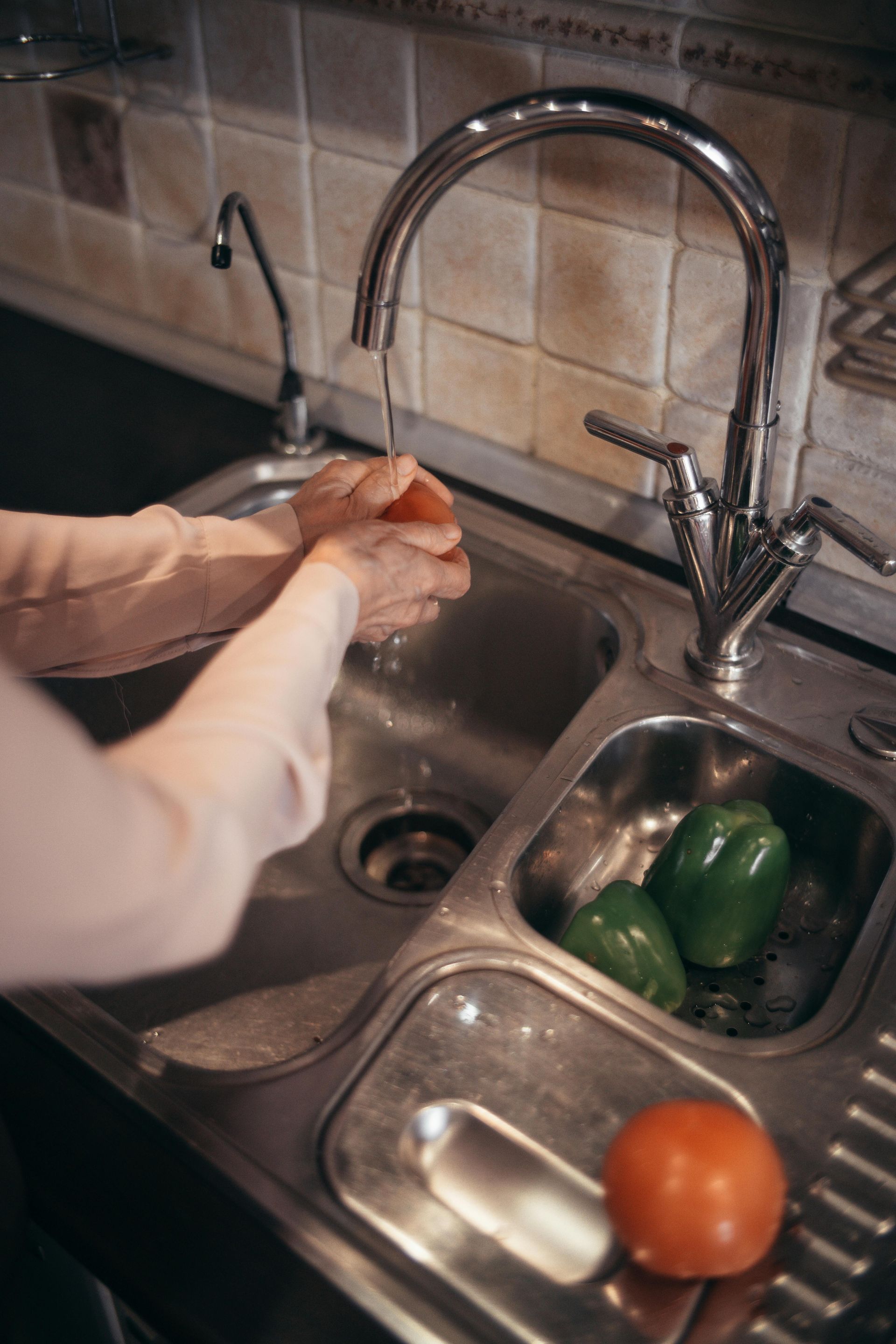Person washing a tomato at a kitchen sink. Green bell peppers in the strainer.