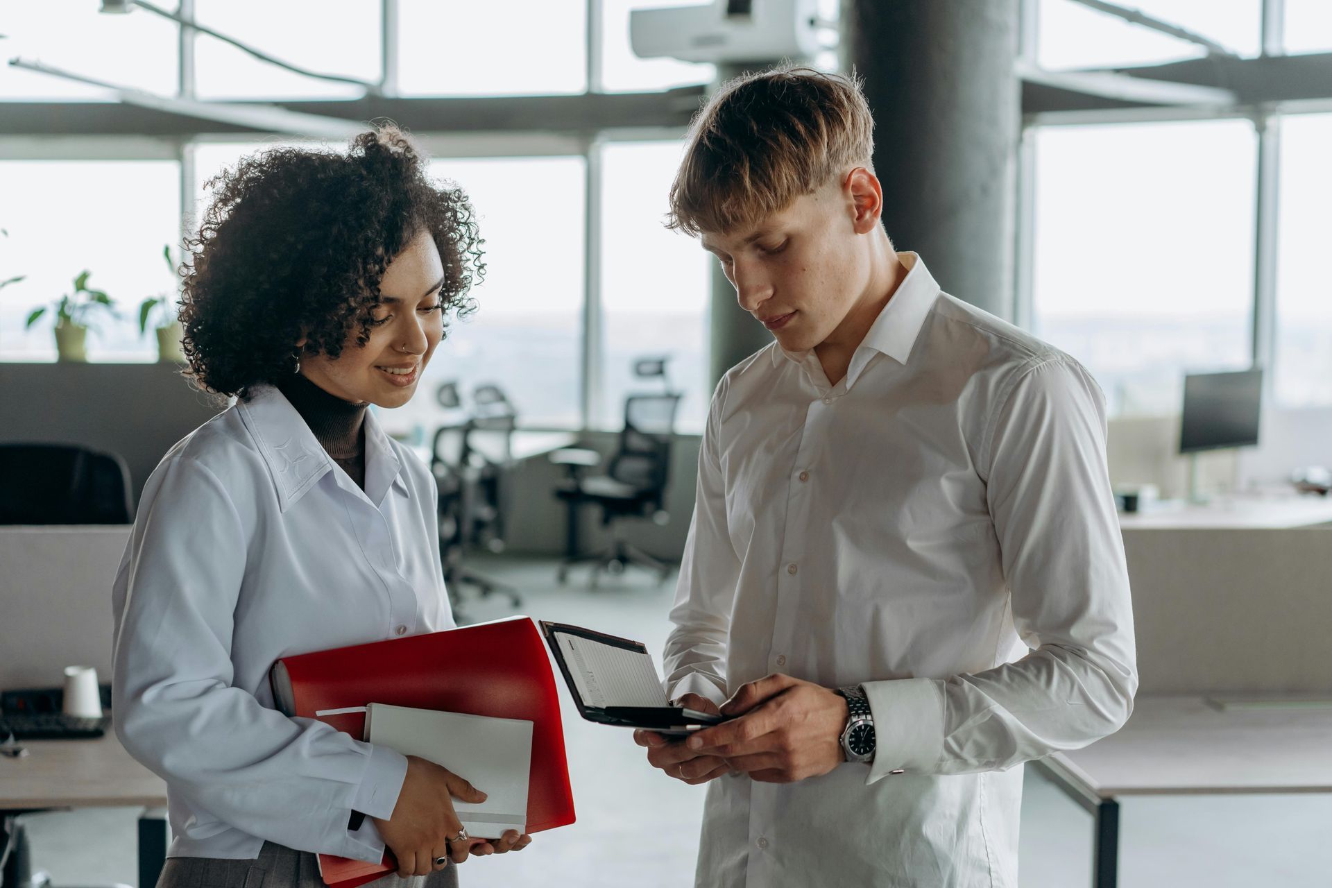 Woman and man in office look at phone. Woman holds folders, both wear white shirts.