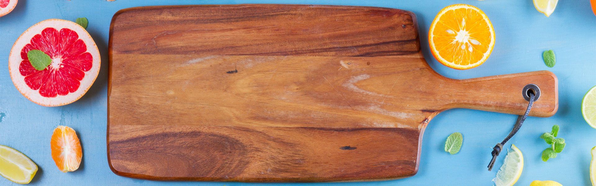 A wooden cutting board surrounded by fruit on a blue table.
