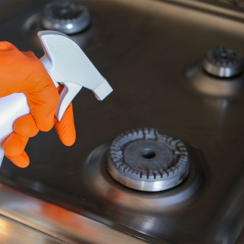 A person wearing orange gloves is cleaning a stove with a spray bottle.
