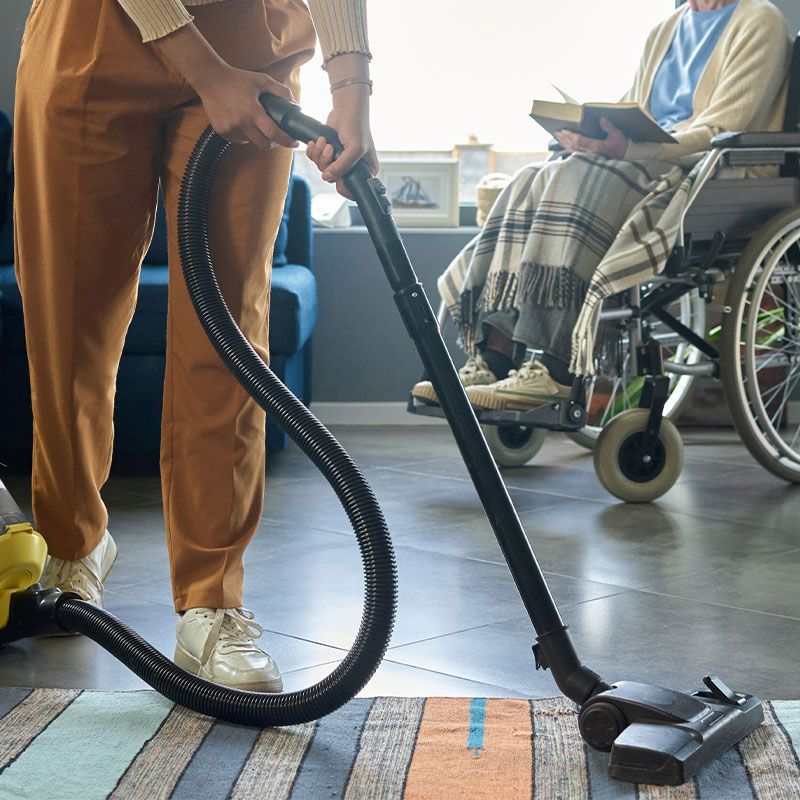 A woman is vacuuming a rug in a living room with an elderly woman in a wheelchair.