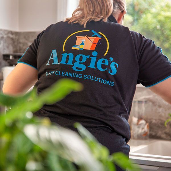 A woman wearing a black angie 's cleaning solutions shirt is standing in a kitchen.