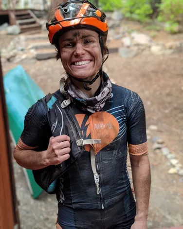 Smiling person with dirt on face wearing bike helmet, cycling gear, and backpack in a wooded setting.