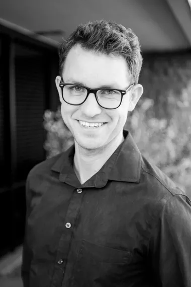 Man with glasses and button-down shirt smiling outdoors.
