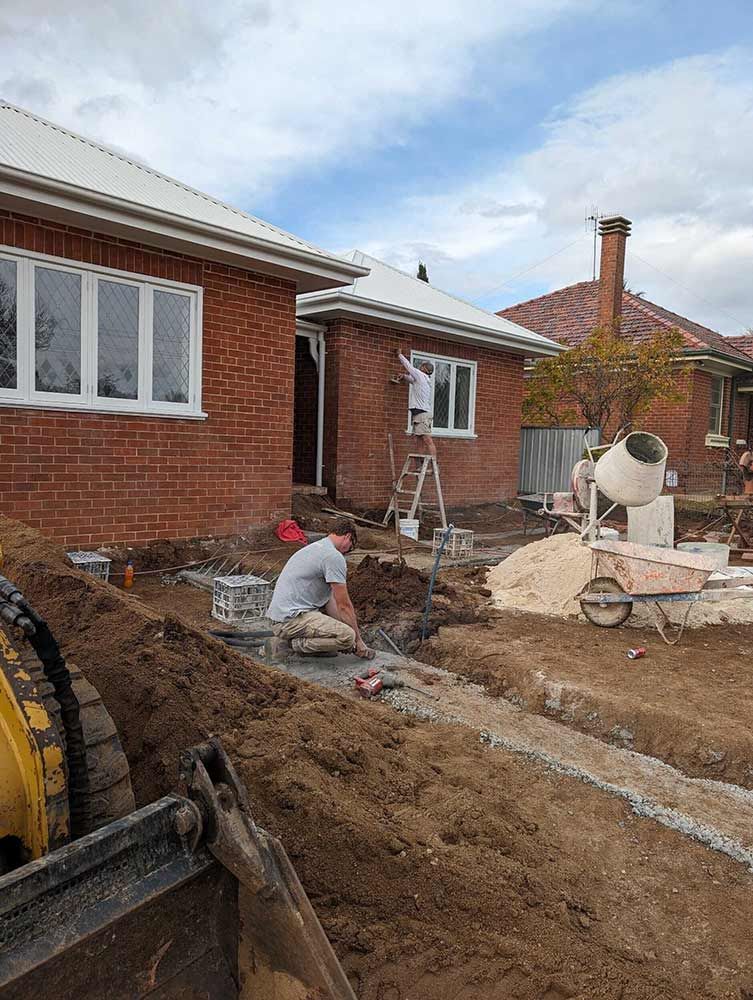 A Man Is Kneeling In The Dirt In Front Of A Brick House — Chandolin Construction In Robin Hill, NSW