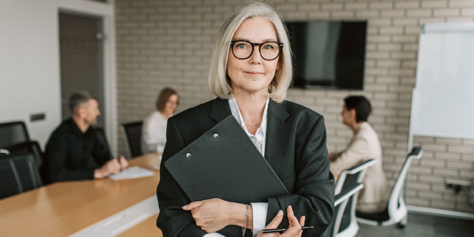 Woman in a suit, holding a clipboard, smiles at the viewer in a meeting room with people working at a table.