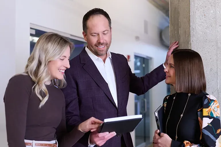 Two women and a man in a modern office space reviewing a tablet together, all smiling during a professional conversation.
