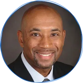 Man in navy blazer smiles in front of a brick wall.