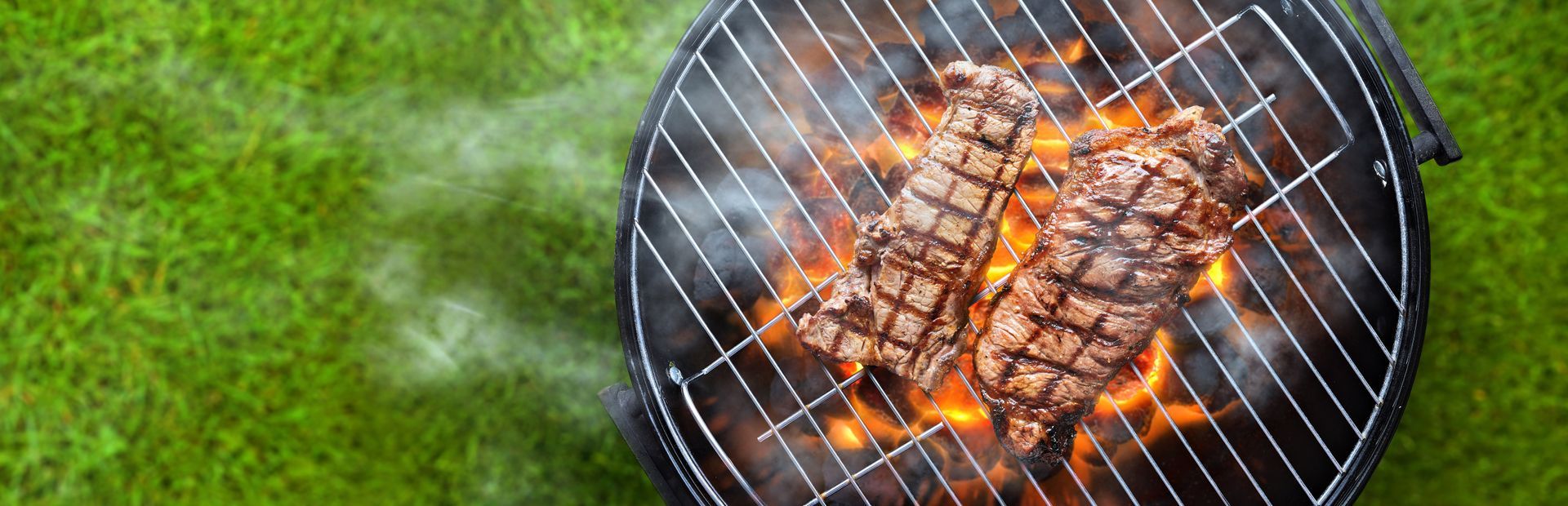 Two steaks grilling over open flames on a round barbecue set on green grass.