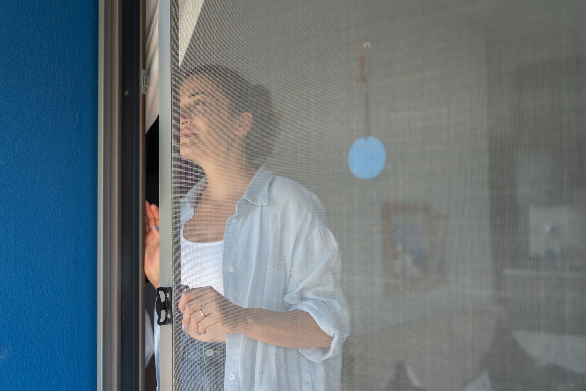 Woman opening a sliding door screen while looking outside, wearing a casual outfit in a bright home.