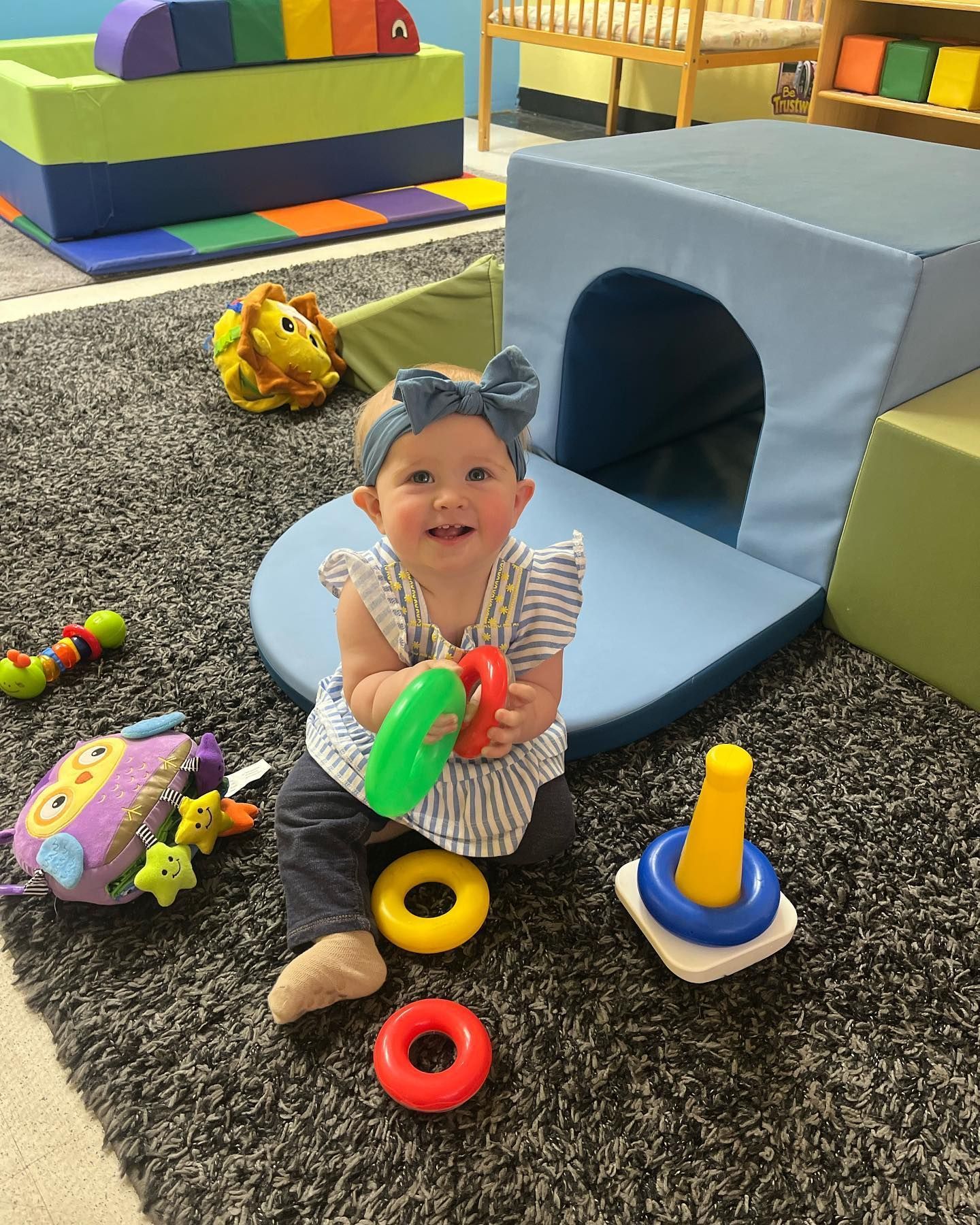 Happy baby holding toys, smiling, in a play area with colorful blocks and a tunnel.
