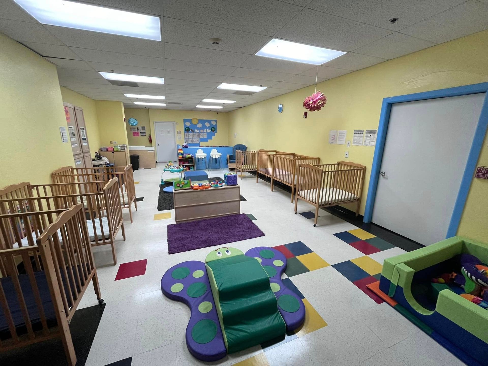 Childcare room with cribs, play equipment, and a colorful floor. Yellow walls and white door.