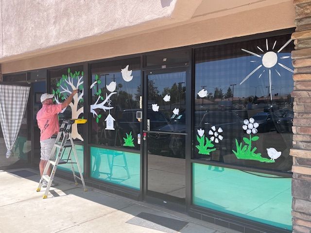 Man on a ladder painting spring-themed decorations on a storefront window.