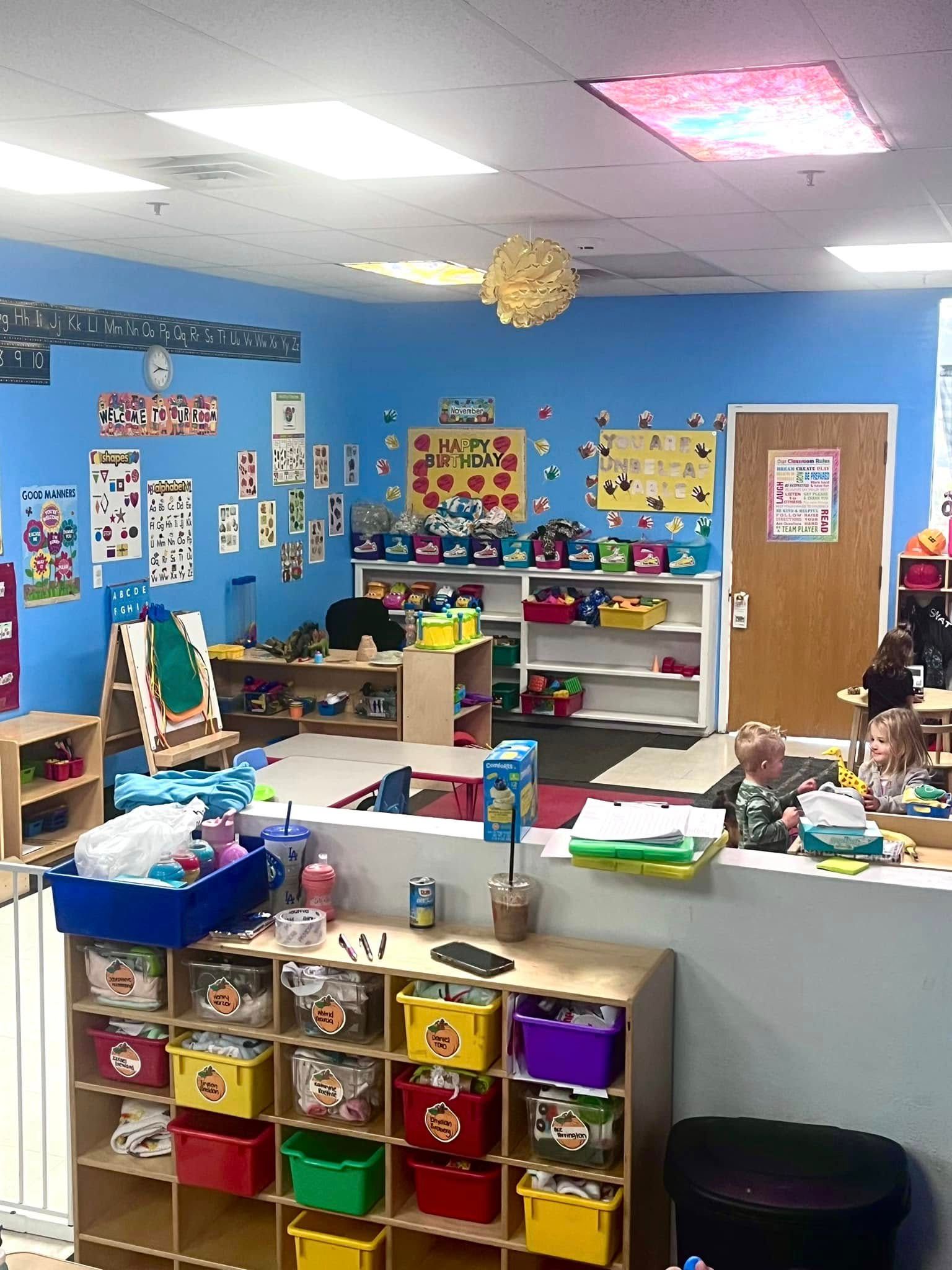 Bright, blue-walled preschool classroom with educational materials, shelves of toys, and cubby storage.