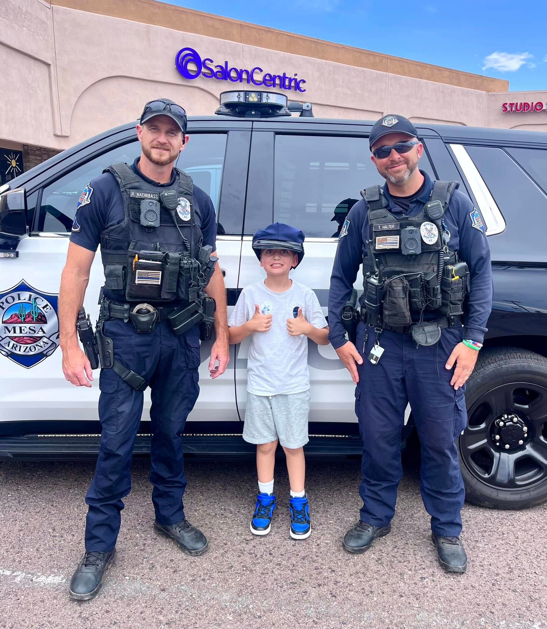 Two police officers and a child pose in front of a police car. The child gives a thumbs up.