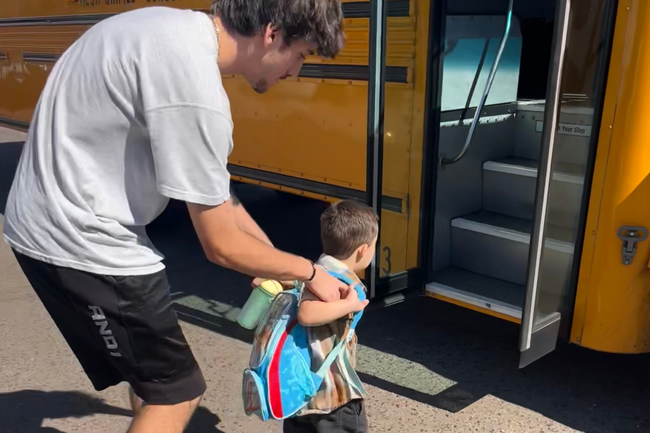 Man helping a child with a backpack board a yellow school bus.