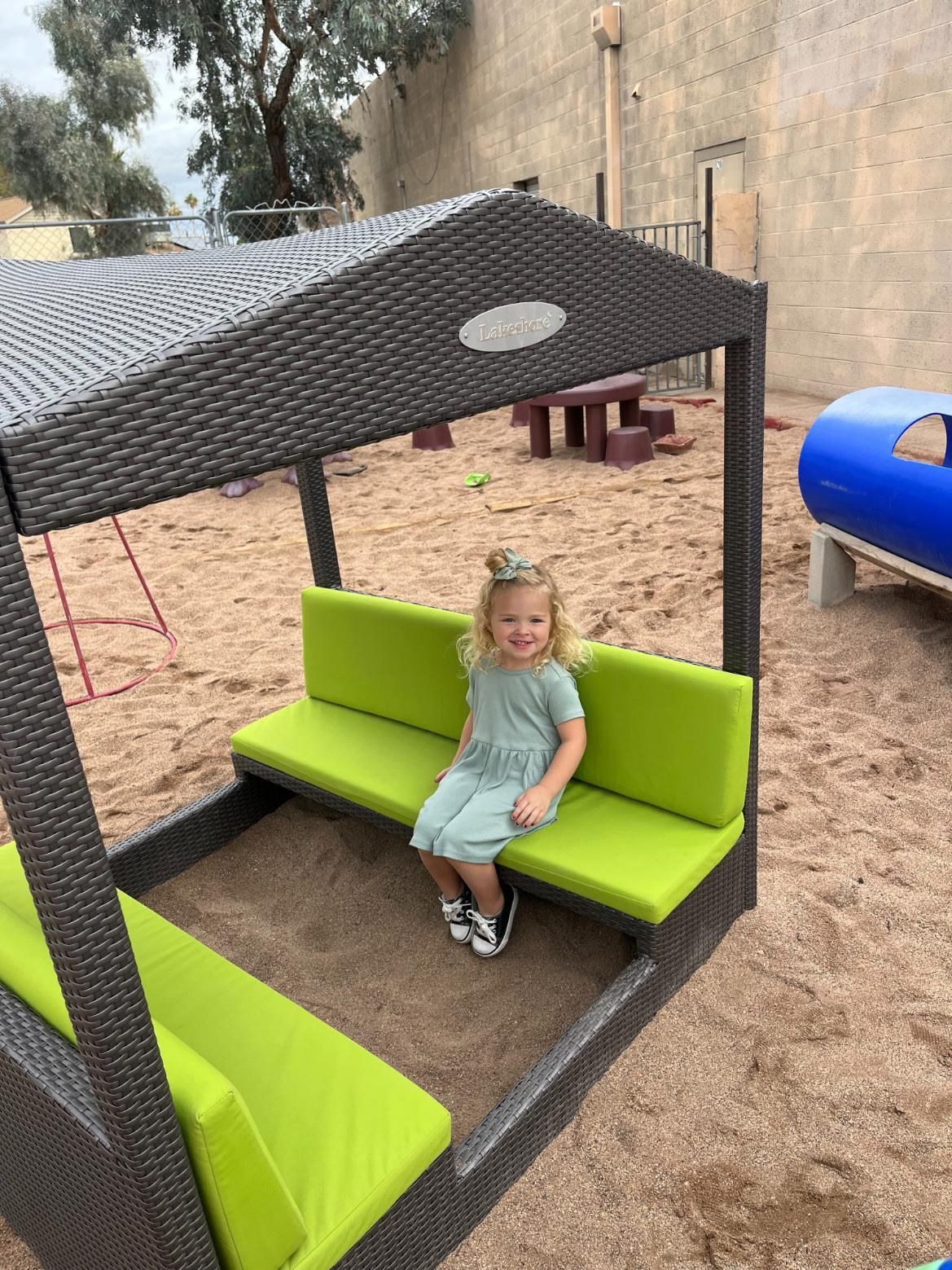 Girl in green dress sits on a sandbox bench with green cushions under a woven structure.