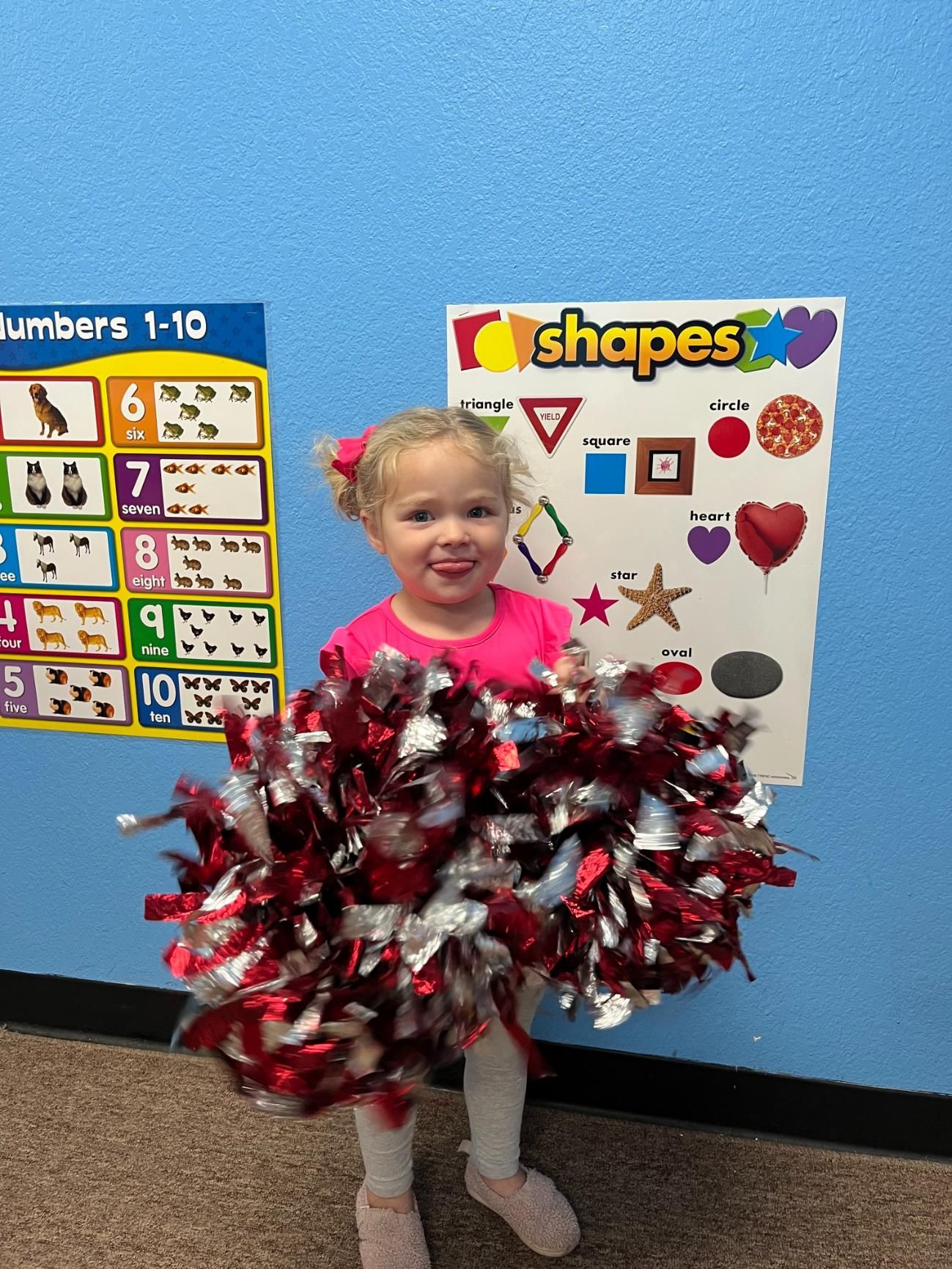 Girl holding red and silver pom-poms, smiling in front of shape and number posters.