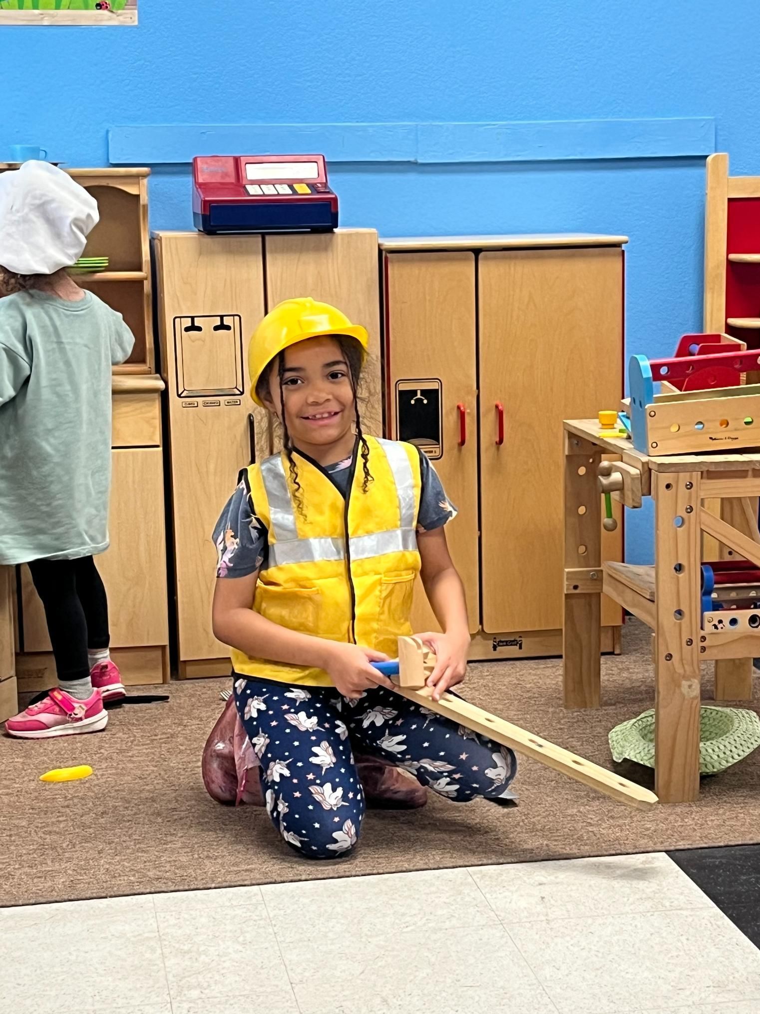 Child in yellow safety vest and helmet playing with toy tools in a play area.