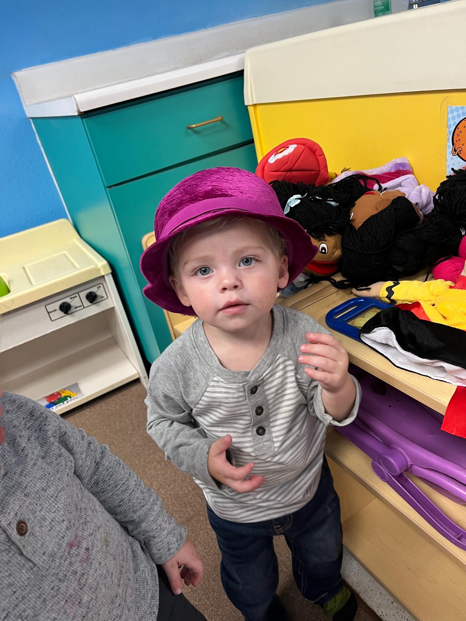Young child wearing a purple hat in a playroom, looking up with a neutral expression.